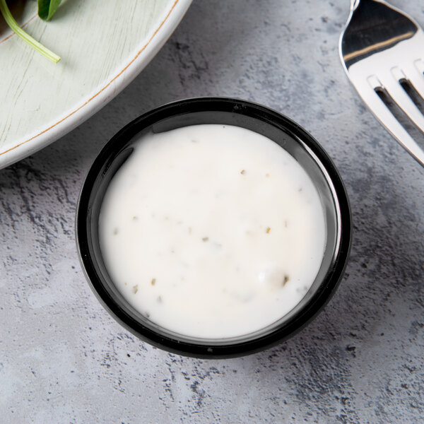 A Tablecraft black melamine ramekin filled with white sauce on a plate with green leaves.