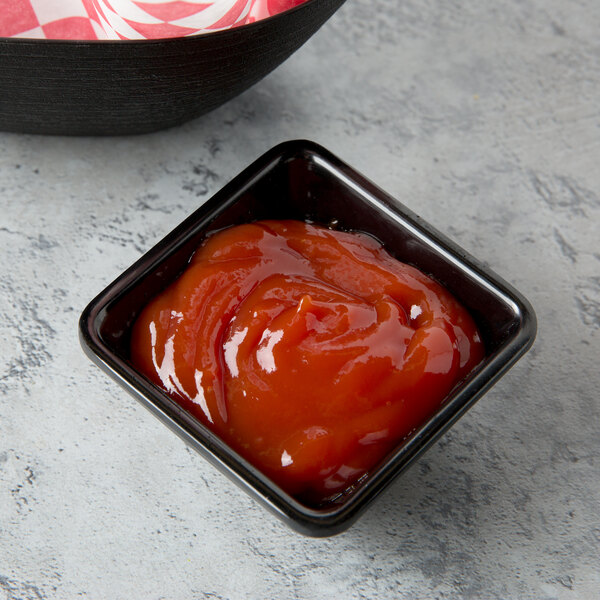 A black square Tablecraft melamine ramekin filled with ketchup on a table.