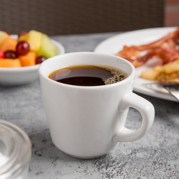 A Libbey Porcelain tall cup of coffee on a table with a plate of food.