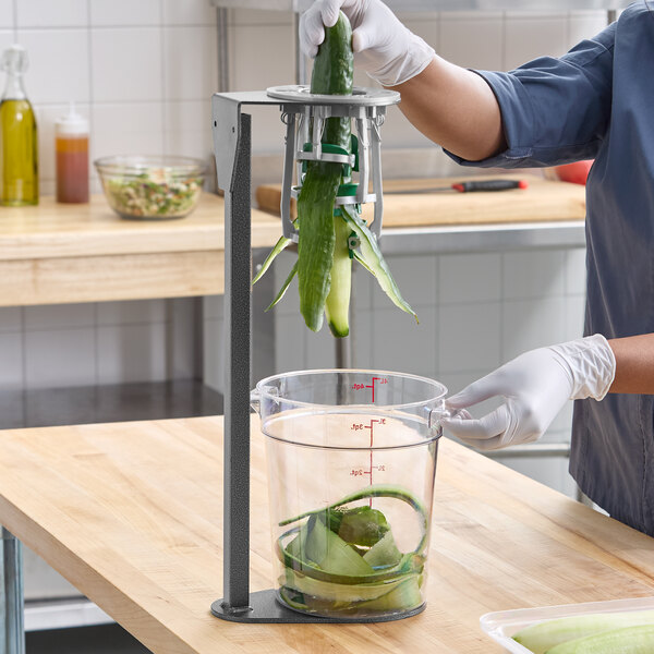A manual upright cucumber peeler being used to peel a cucumber over a clear container.