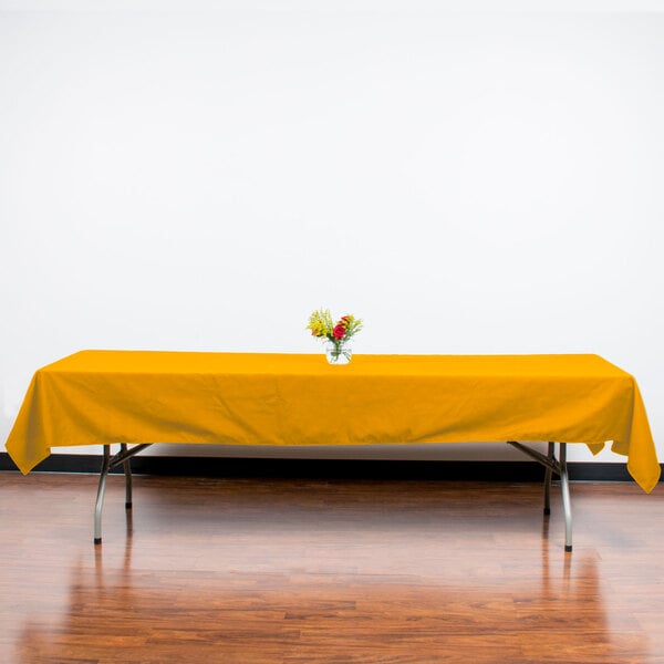 A table with a yellow Intedge rectangular tablecloth on it with a vase of flowers.