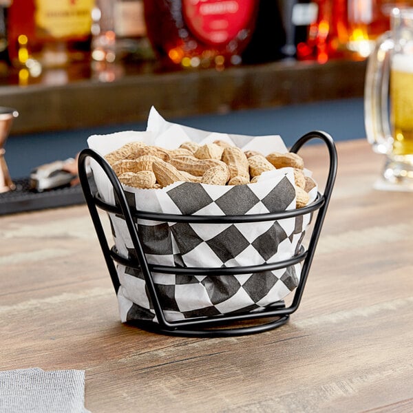 A black Clipper Mill round wire basket filled with peanuts on a table.