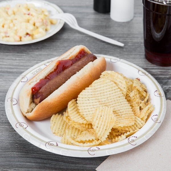 Hot dog and waffle chips on a paper plate with a plate of macaroni salad in the background