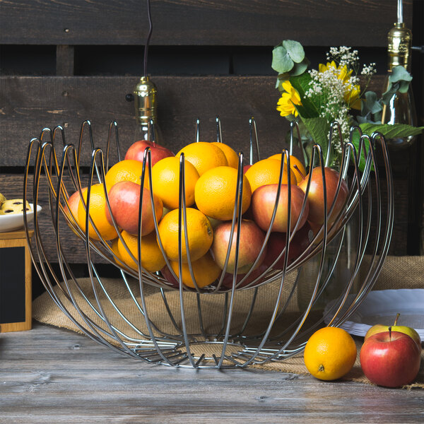 A Clipper Mill chrome plated iron wire basket filled with apples and oranges on a table.