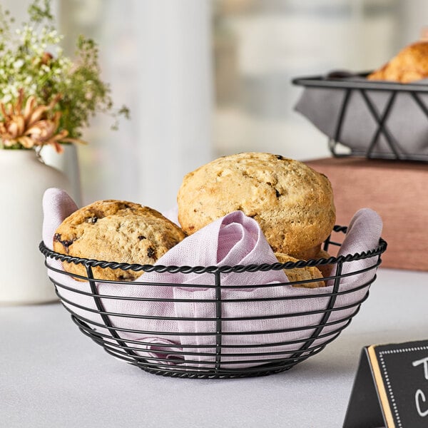 A black powder coated iron round wire basket with muffins and a napkin inside on a table.