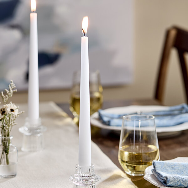 A white 10-inch taper candle burning in a clear candle holder on a dining table.
