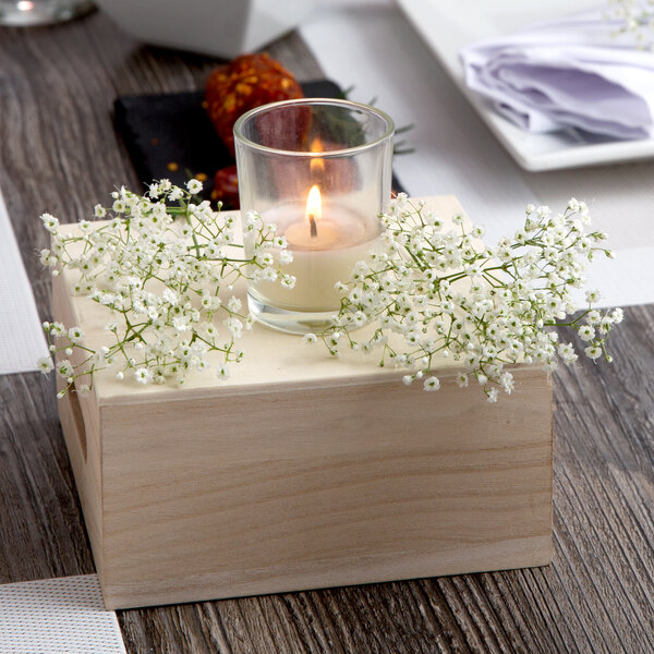 A Leola clear glass wax filled votive candle with a flame on a table with flowers.