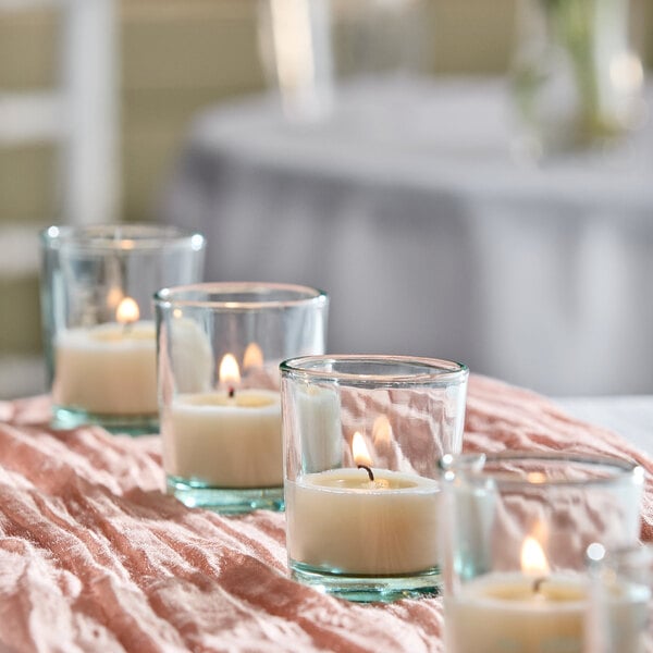 A set of clear glass votive holders filled with white wax candles, each lit and arranged in a row on a textured pink cloth.