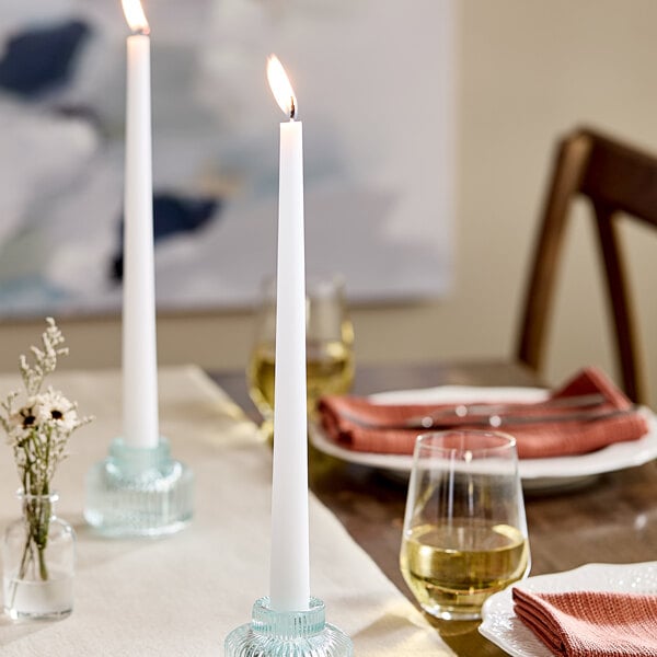 A white 12-inch taper candle burning in a glass candle holder on a set dining table.