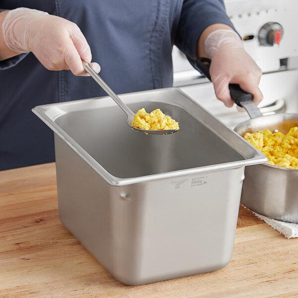 A hand using a spoon to serve scrambled eggs from a Vollrath stainless steel container.