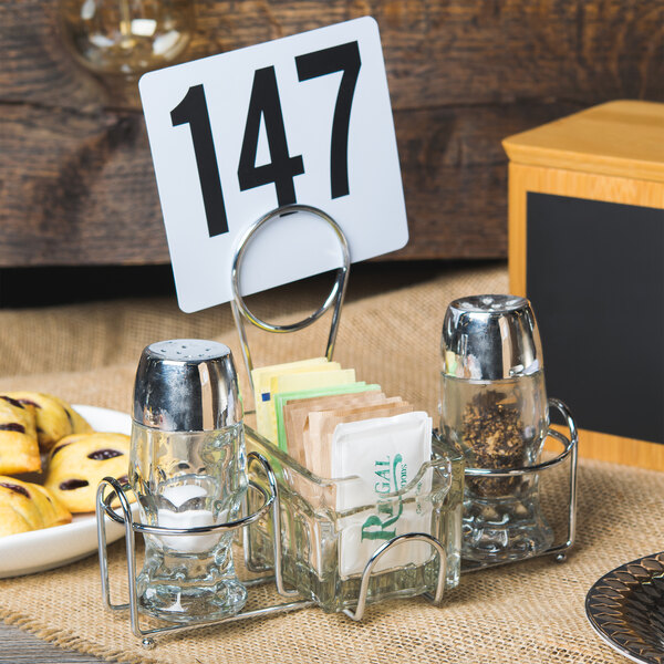 A Clipper Mill 2-compartment condiment caddy with a menu holder on a table with a clear glass container of condiment packets.