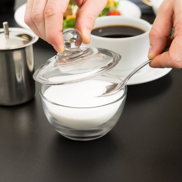 A person pouring sugar from a spoon into an Anchor Hocking glass sugar bowl.