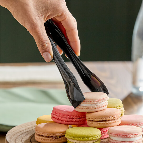A pair of black disposable plastic tongs being used to pick up a macaron from a stack of assorted macarons.