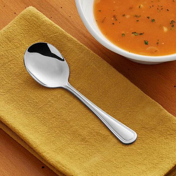 An Acopa stainless steel bouillon spoon on a napkin next to a bowl of soup.