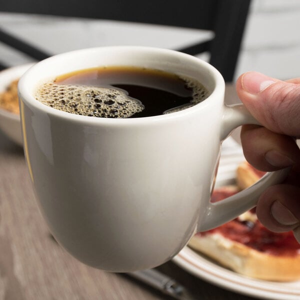 A hand holding a Tuxton white china coffee mug filled with coffee.