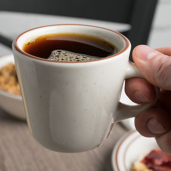 A hand holding a Tuxton Bahamas brown speckle tall china cup of coffee.