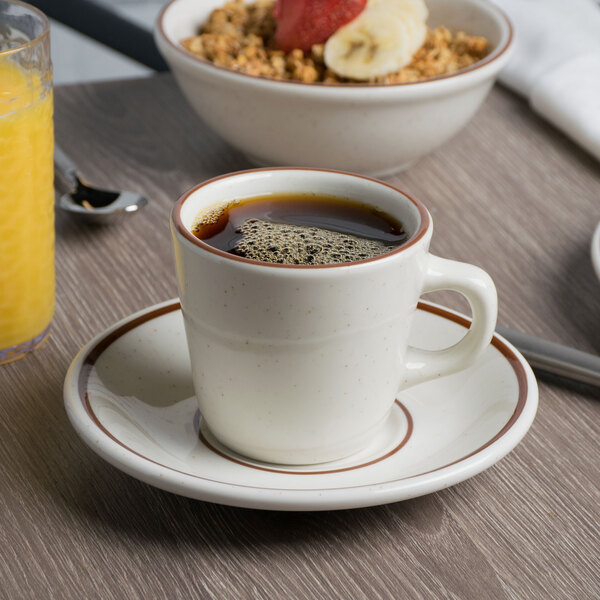 A Tuxton Bahamas brown speckle tall cup of coffee on a saucer on a table with a bowl of cereal and fruit.