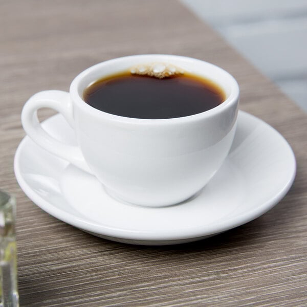 A Libbey Basics bright white porcelain espresso cup on a saucer with a cup of coffee on a table.