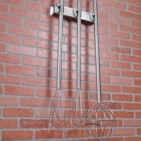 A Matfer Bourgeat utensil holder rack with three metal whisks hanging on a metal pole against a brick wall.