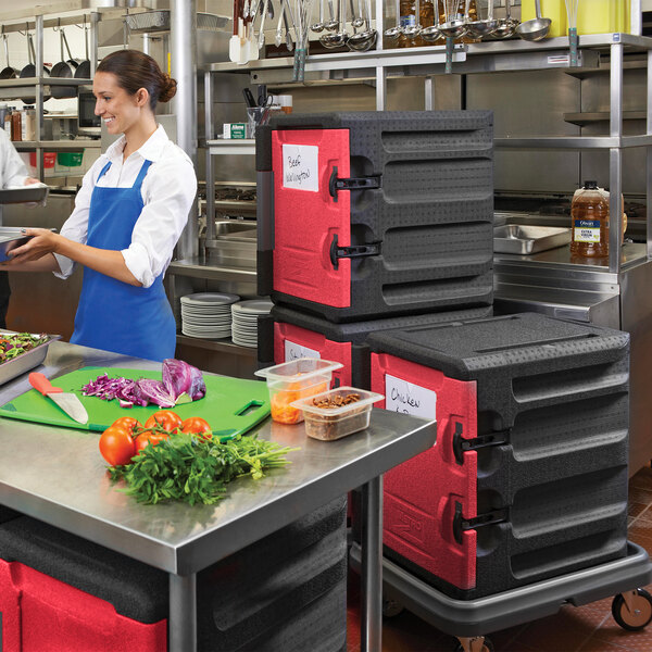 A woman in a blue apron standing in a kitchen using a black and red Metro Mightylite food pan carrier.