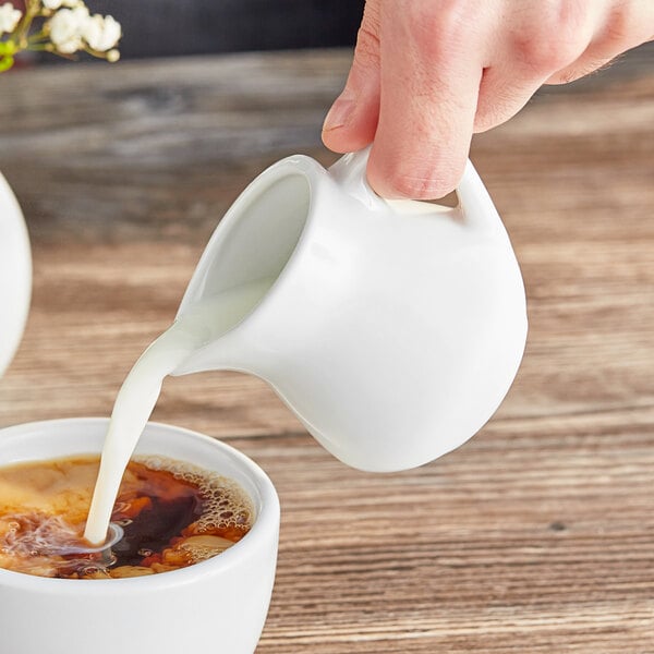 A hand pouring milk from an Acopa white creamer into a cup of coffee.
