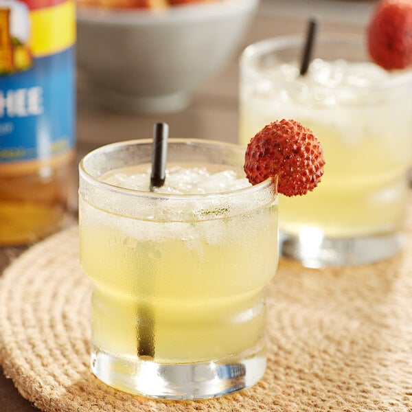 A glass of yellow liquid with a straw and a lychee fruit on the rim.