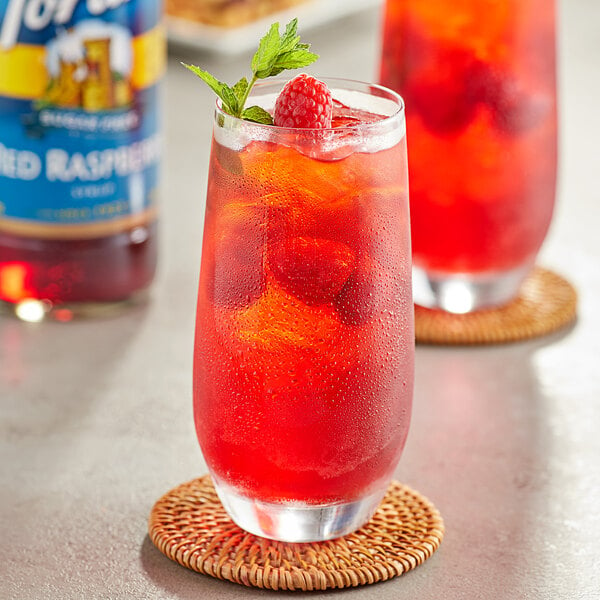 A glass of red raspberry drink garnished with a raspberry next to a bottle of Torani Sugar-Free Red Raspberry Flavoring Syrup.