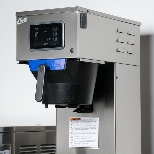 A Curtis combination tea and coffee brewer on a counter in a professional kitchen with a black plastic container and blue and black handles.