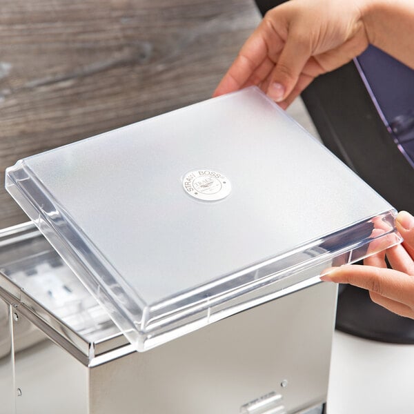 A hand opening a box with a clear plastic lid with a logo on a piece of paper.