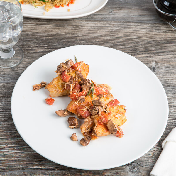 A Schonwald round porcelain coupe plate with pasta, meat, mushrooms, and tomatoes on a wooden table.