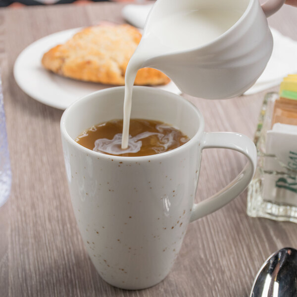 A person pouring milk from a white pitcher into a white Schonwald porcelain mug of coffee.