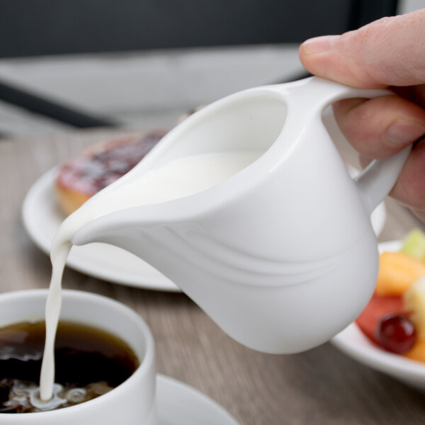 A person pouring milk from a white Schonwald creamer into a cup of coffee.