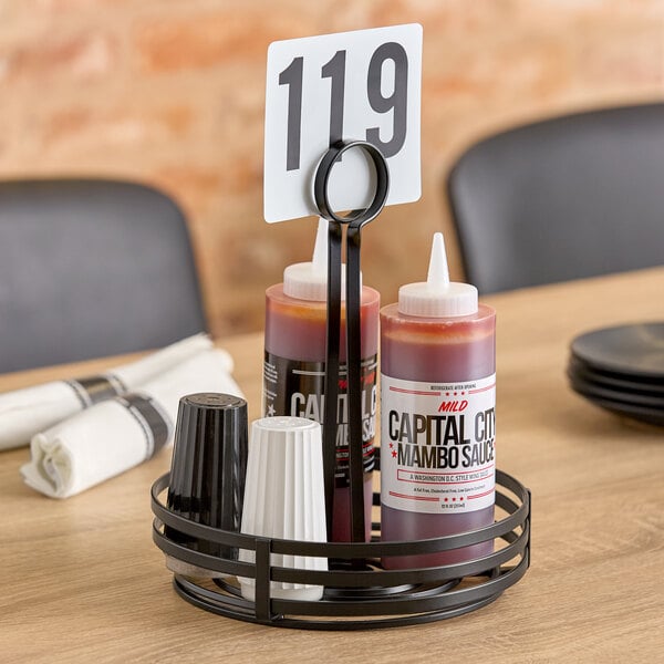 A black wrought iron round condiment caddy with a card holder, holding sauce bottles and shakers.