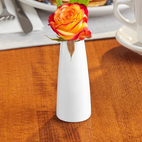 A small, white porcelain bud vase holding a single orange and yellow rose.