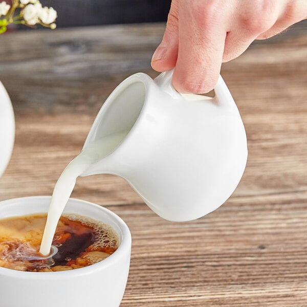 A hand pouring cream from an Acopa white creamer into a cup of coffee.