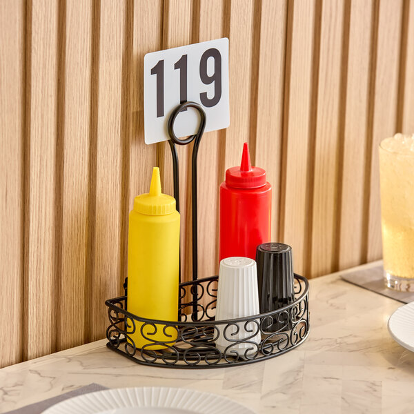 A black wrought iron half round condiment caddy with a card holder, holding yellow and red squeeze bottles and salt and pepper shakers.