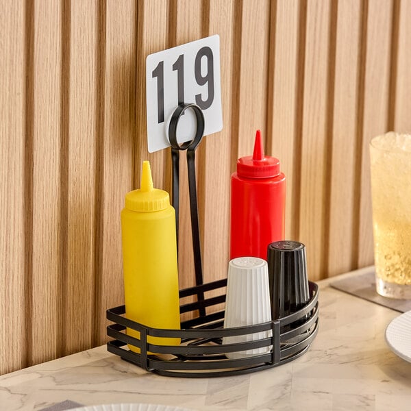 A black wrought iron condiment caddy with a card holder, containing squeeze bottles and shakers, placed on a table.
