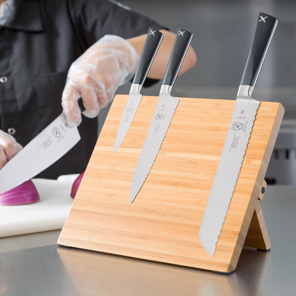 A person cutting a vegetable with a Mercer Culinary ZüM® knife on a bamboo cutting board.