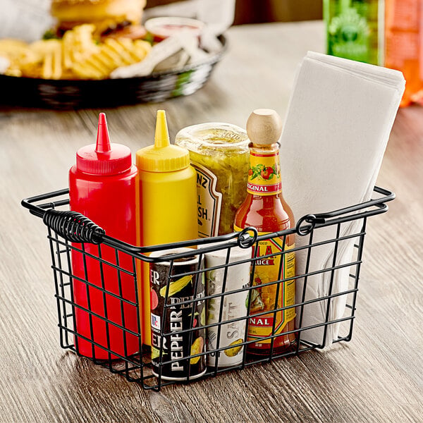 A black wire tabletop market basket with condiments including a jar of pickles and a bottle of mustard.