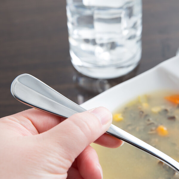 A hand holding a Libbey stainless steel round soup spoon over a bowl of soup.