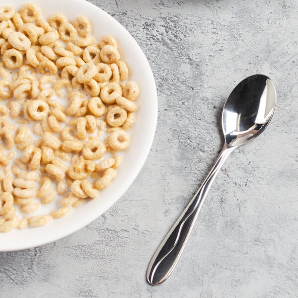 A bowl of cereal with milk and a Libbey stainless steel teaspoon on a table.