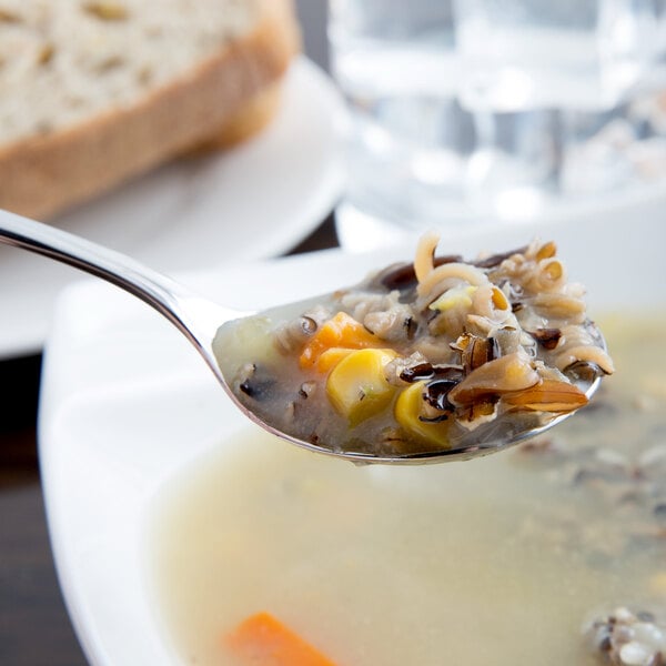 A Libbey stainless steel bouillon spoon filled with soup next to a piece of bread.