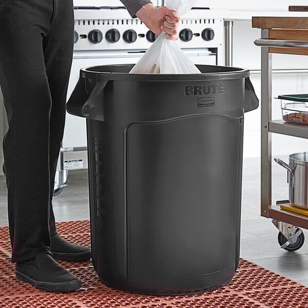 A man in a professional kitchen putting a plastic bag into a black Rubbermaid BRUTE trash can.