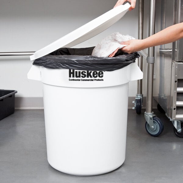 A person putting a black bag into a white Continental Huskee trash can.