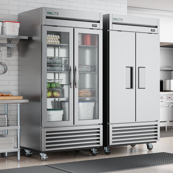 A room in a professional kitchen with two True 2 section glass door reach-in refrigerators.