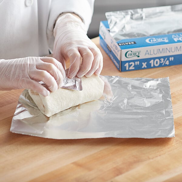 A person in gloves is wrapping food using a box of Choice interfolded foil sheets.