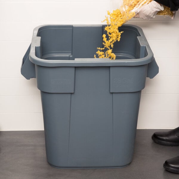 A person pouring food into a Rubbermaid grey square trash can.