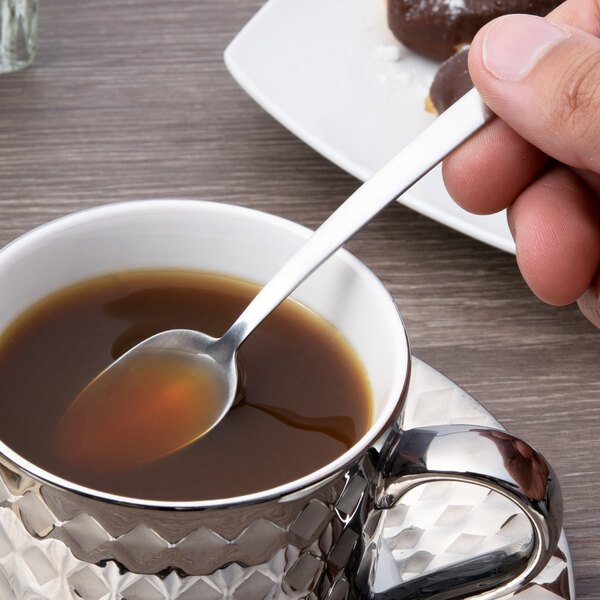 A hand holding an Arcoroc stainless steel demitasse spoon over a cup of liquid.