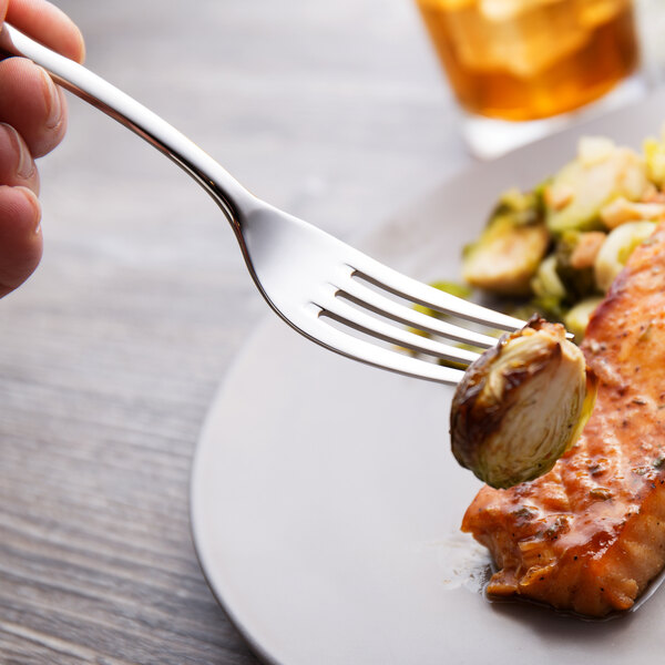 A hand holding a Chef & Sommelier stainless steel dinner fork over a plate of meat with brussels sprouts.