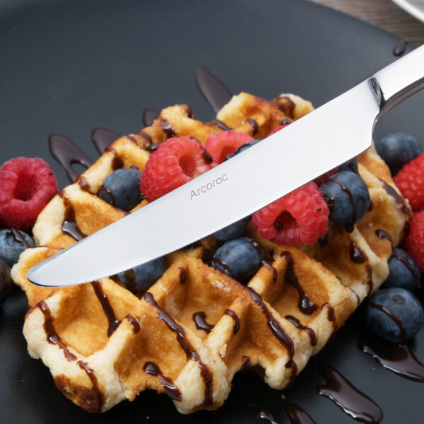 An Arcoroc stainless steel dessert knife cutting into a waffle topped with berries and chocolate sauce.